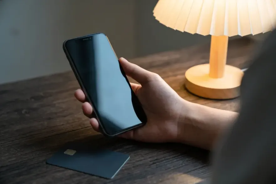 Hand holding a phone with the screen switched off beside a plain bank card on a wooden desk